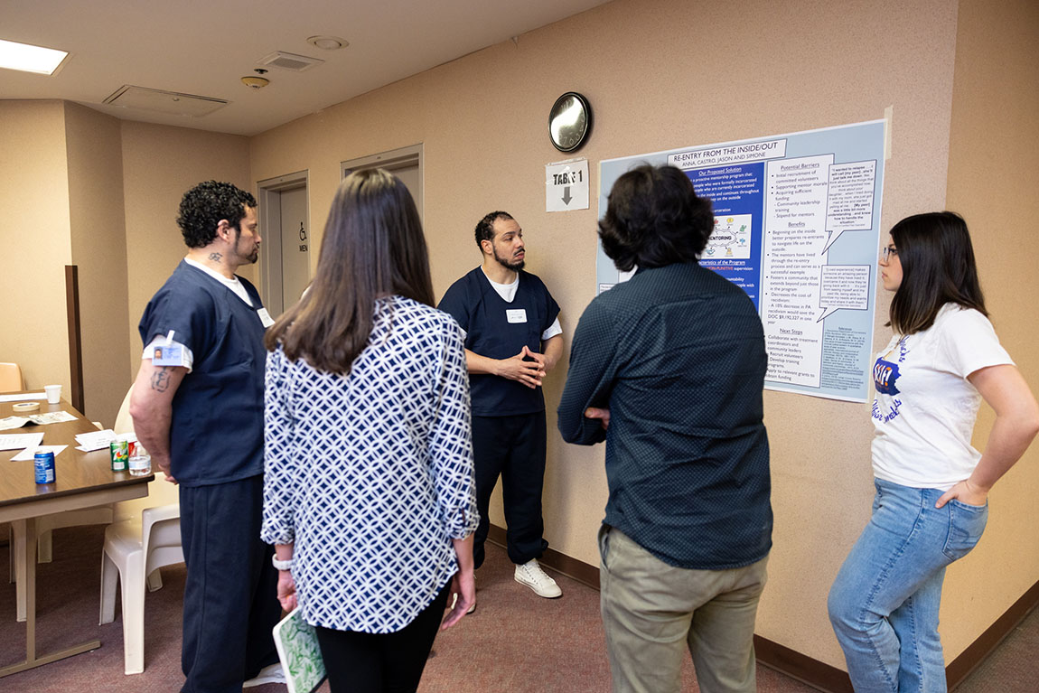 A group gathers around a poster as a man with a beard wearing a navy blue prison uniform explains the poster's contents